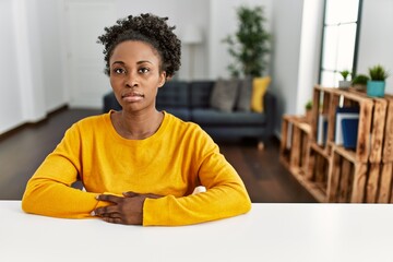 Sticker - Young african american woman wearing casual clothes sitting on the table at home relaxed with serious expression on face. simple and natural looking at the camera.