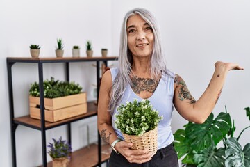 Poster - Middle age grey-haired woman holding green plant pot at home smiling cheerful presenting and pointing with palm of hand looking at the camera.