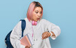 Hispanic woman with pink hair wearing student backpack and headphones looking at the watch time worried, afraid of getting late