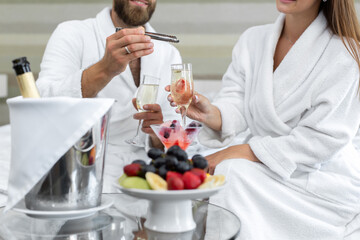 Man puts berries in a glass of sparkling wine to his woman in a hotel in bed.