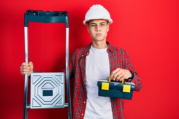 Wall Mural - Young hispanic man holding tools box wearing hardhat by construction stairs puffing cheeks with funny face. mouth inflated with air, catching air.