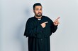 Handsome hispanic man with beard wearing catholic priest robe pointing aside worried and nervous with both hands, concerned and surprised expression