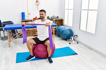 Canvas Print - Young hispanic man patient smiling confident having rehab session using fit ball and elastic band at clinic