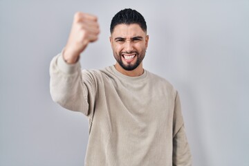 Canvas Print - Young handsome man standing over isolated background angry and mad raising fist frustrated and furious while shouting with anger. rage and aggressive concept.