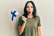 Young hispanic girl holding finland flag scared and amazed with open mouth for surprise, disbelief face
