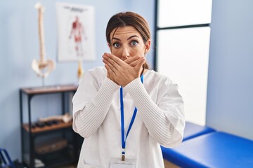 Wall Mural - Young brunette woman working at pain recovery clinic shocked covering mouth with hands for mistake. secret concept.