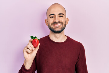 Canvas Print - Young bald man holding strawberry looking positive and happy standing and smiling with a confident smile showing teeth