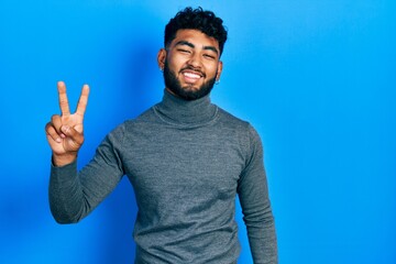 Poster - Arab man with beard wearing turtleneck sweater smiling looking to the camera showing fingers doing victory sign. number two.