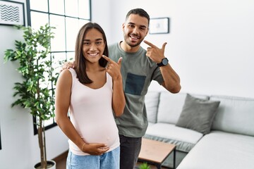 Sticker - Young interracial couple expecting a baby, touching pregnant belly smiling cheerful showing and pointing with fingers teeth and mouth. dental health concept.