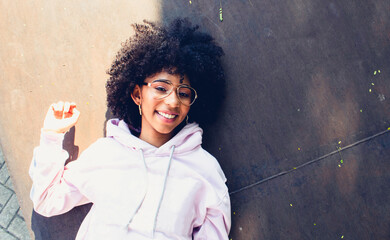 Wall Mural - portrait of a smiling afro woman lying on the floor isolated with glasses and sweatshirt having fun on a sunny day, with color background