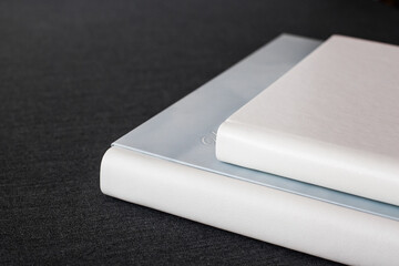 Two white stylish photobooks with leather covers, different sizes, lie on a dark gray surface in a room.