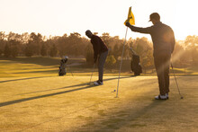 African American Young Man Playing Golf With Caucasian Friend Holding Flag At Golf Course At Sunset