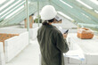 © sonyachny - Young female architect with tablet checking blueprints against wooden roof framing of modern farmhouse. Stylish woman engineer in hard hat looking at digital plans at construction site