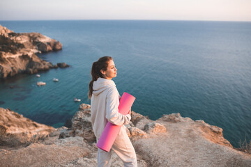 Wall Mural - Happy smiling woman holding pink yoga mat wear sportswear hoodie and pants standing on top of rock over sea side background. Pretty adult girl practice fitness at open air outside. Healthy lifestyle.