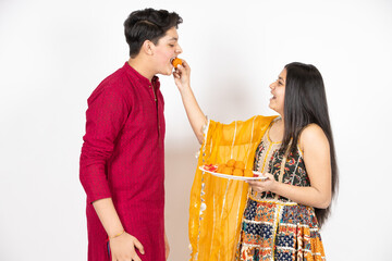 Young Indian brother and sister celebrating Diwali, rakshabandhan or bhai dooj festival isolated on white studio background. enjoying sweet laddo,