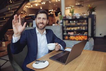 Wall Mural - Caucasian handsome man with coffee waving hand, looking at window at laptop in cafe. Meeting concept