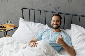 Wall Mural - Young smiling happy fun man wear casual blue t-shirt lying in bed show thumb up gesture look camera rest relax spend time in bedroom lounge home in own room house wake up dream. Real estate concept.