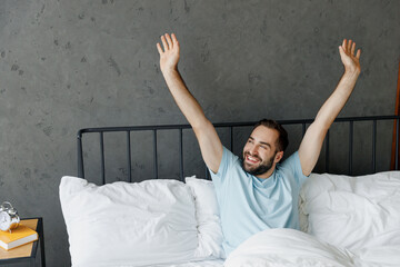 Wall Mural - Young satisfied smilng happy fun man wear casual blue t-shirt lying in bed look aside stretch hands rest relax spend time in bedroom lounge home in own room house wake up dream. Real estate concept.