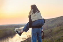 A Beautiful Young Girl Sat On The Back Of A Guy, They Run And Have Fun While Relaxing Outside The City On A Walk In The Rays Of The Setting Sun, Below The River