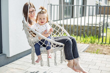 Happy Mom And Little Baby Girl Are Swinging On Swing Chair In Backyard Of House.