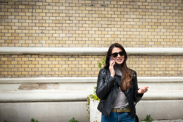 Young business woman talking on smartphone outdoor. Attractive girl in leather jacket having conversation with her friends waiting for the to go on the rock music concert.