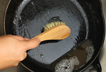 Close up A hand holding bamboo cleaning brush scrub and washing the dirty cast iron pan with a pattern of grease and oil stains.