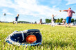 Youth sized glove with a baseball inside sitting on a grass field with players in the background