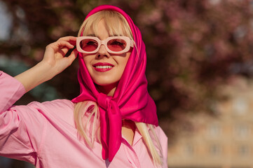 Fashionable happy smiling woman wearing trendy pink rectangle sunglasses, fuchsia color headscarf,  classic shirt, posing in street of city. Copy, empty space for text