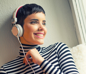 Beautiful young woman listening music through earphones while sitting beside window in her room at home