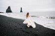Girl in old old-styled wedding dress are running on the Black Sand beach in Vic, Iceland with rock islands on background.
