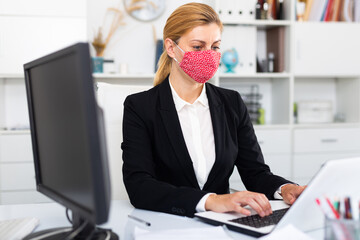 Wall Mural - Young businesswoman in colored medical mask working with laptop and documents in office