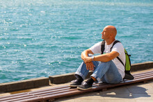 Traveler Sits On Waterfront, Middle-aged Man Tourist With Backpack, Sits On Wooden Deck And Looks At Blue Sea On A Sunny Day, A Caucasian Man Alone On The Pier Rests And Meditates
