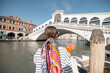 Young woman enjoying summer cocktail on the background of famous Rialto bridge in Venice. Concept of happy vacation and leisure time in Italy. Standing back with italian alcohol drink spritz Aperol