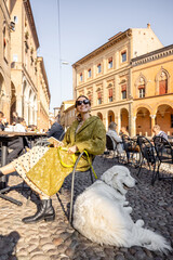 Stylish woman sitting with her white dog at outdoor cafe in the old town of Bologna city. Italian measured lifestyle and street fashion concept. Idea of traveling Italy