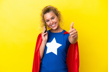 Wall Mural - Super Hero woman isolated on yellow background keeping a conversation with the mobile while doing thumbs up