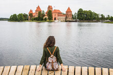Trakai Castle In Lithuania. Young Woman (back View) Sitting On The Dock And Looking At The Castle, Trakai, Lithuania.