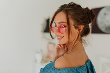 Close-up Photo In Profile Of Fair-skinned Young Girl With Tanned Face On Blur Light Background. Brunette Woman With Wonderful Smile Wearing Round Pink Glasses And Denim T-shirt
