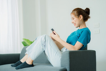 Portrait of a beautiful Asian woman using a smartphone on the sofa in the living room in the bedroom. A happy woman relaxing at home sitting on a sofa using a mobile phone.