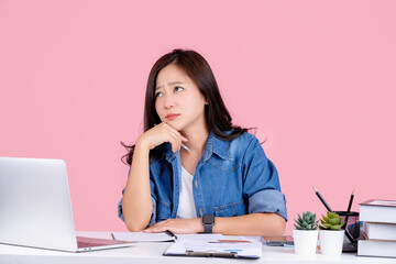 Young dreamy asian woman looking up thinking searching new idea holding pen and sit work at white desk with pc laptop isolated on pastel pink background.