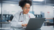 © Gorodenkoff - Hospital Doctors Office: Portrait of Young Promising Black Physician Works on Laptop Computer. Female Medical Professional in White Lab Coat Looking for Patient Treatment While Sitting at Her Desk