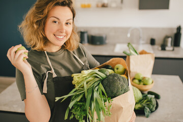 Young woman standing on the kitchen, preparing to cook smoothie using fresh green vegetables and fruits.