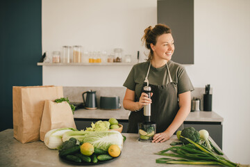 Young woman making smoothie at the kitchen, using blender and fresh fruits and vegetables.