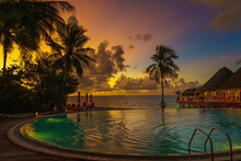 Luxury Sunset Over Infinity Pool In A Summer Beachfront Hotel Resort At Tropical Landscape. Tranquil Beach Holiday Vacation Background Mood. Amazing Island Sunset Beach View, Palms Swimming Pool