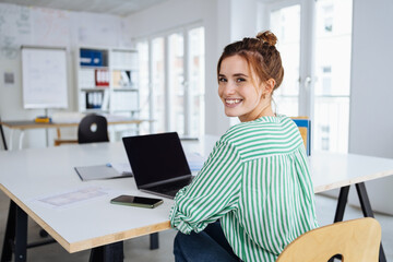 Wall Mural - Young businesswoman sitting in office in front of laptop looking at camera