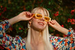 Spring, summer fashion conception: joyful blonde woman wearing trendy yellow rectangle sunglasses posing near blooming tree