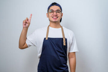 Asian barista man wearing apron and cap showing and pointing up with fingers number one while smiling confident and happy