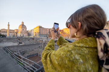 Wall Mural - Happy woman taking photo of Roman forum, while traveling in Rome. Style caucasian woman in coat and shawl in hair having fun traveling italian landmarks