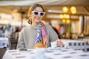 Wall Mural - Woman having breakfast with croissant and coffee at traditional italian cafe outdoor. Woman wearing sunglasses and shawl in hair. Concept of italian lifestyle and travel