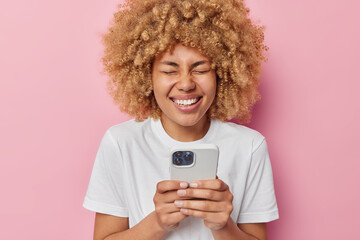 Wall Mural - Joyful curly haired woman types text messages surfs internet and laughs sincerely reads funny content in internet giggles positively dressed in white casual t shirt isolated over pink background.