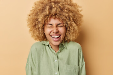 Overjoyed curly haired European woman laughs gladfully keeps eyes closed dressed in casual shirt laughs positively isolated over brown background has fun. People and sincere emotios concept.